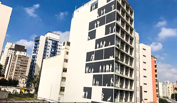 Edifício alto e branco em uma rua de São Paulo, com painéis pretos na fachada decorados com silhuetas brancas de pessoas em diferentes atividades. Ao redor, há outros prédios residenciais, sob um céu azul com nuvens.