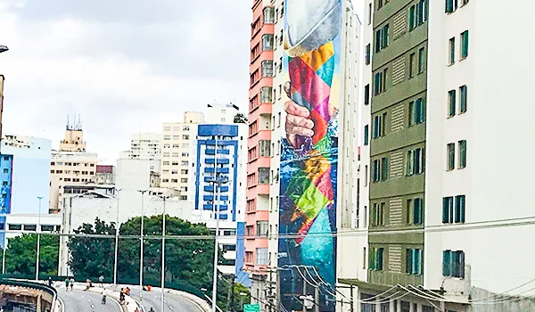 Vista urbana de São Paulo, com edifícios altos e coloridos, incluindo um prédio com um grande mural de arte urbana que retrata mãos e formas geométricas vibrantes. Ao fundo, há outros prédios residenciais e comerciais, árvores e uma via elevada com veículos e pedestres.