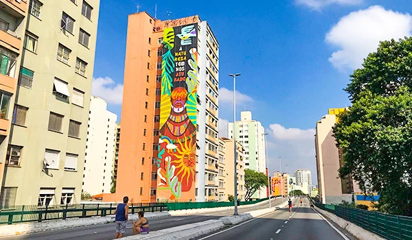 Vista de uma avenida urbana em São Paulo, com prédios residenciais e um edifício laranja decorado com um grande mural colorido de arte urbana. A via asfaltada se estende entre os edifícios, com pedestres caminhando na calçada, árvores ao lado direito e céu azul com nuvens ao fundo.