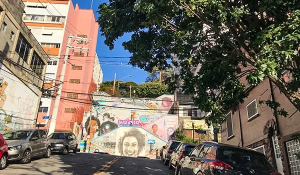 Rua em aclive em São Paulo, com carros estacionados nas laterais, prédios residenciais e comerciais e árvores que oferecem sombra. Ao fundo, um muro exibe grafites coloridos, incluindo um retrato de Marielle Franco.