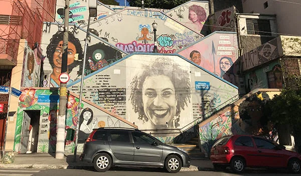Mural colorido em uma fachada urbana, com diversos grafites e retratos de pessoas, incluindo um grande rosto sorridente no centro da parede. Ao redor, há desenhos, frases, assinaturas e elementos artísticos em diferentes cores. Na parte inferior, dois carros estacionados em frente ao mural, postes e árvores completam a cena.