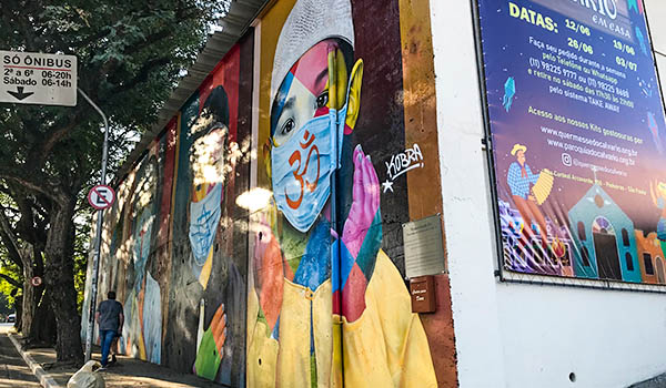 Muro de uma igreja coberto por grafites coloridos, com destaque para um rosto usando máscara de proteção e mãos em tons vibrantes. Ao lado, há um cartaz informativo sobre eventos, enquanto pedestres caminham pela calçada arborizada de uma rua de São Paulo.