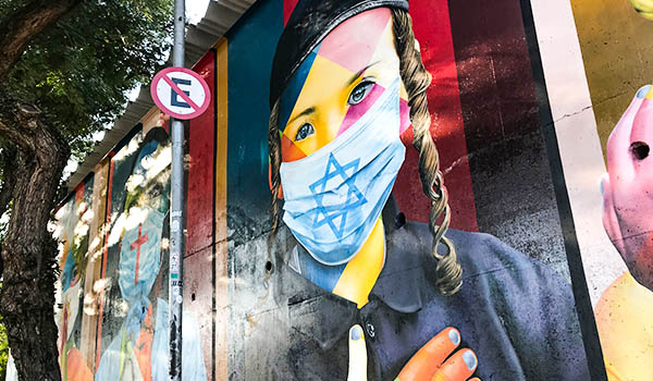 Mural de grafite em uma parede urbana retratando uma mulher com tranças e máscara azul-clara estampada com a Estrela de Davi, em meio a formas geométricas e cores vibrantes. À esquerda, há uma árvore e uma placa de trânsito de proibido estacionar.