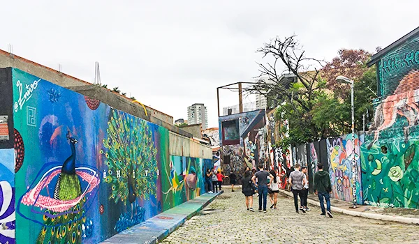 Beco do Batman, em São Paulo, com vielas de paralelepípedos cercadas por muros cobertos de grafites coloridos, incluindo um pavão e desenhos artísticos. Visitantes caminham pelo local ao ar livre, cercado por árvores e construções urbanas, sob um céu nublado.