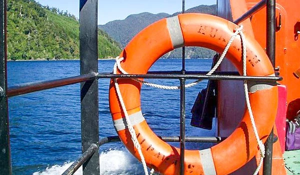 Salva-vidas laranja pendurado na grade de um barco em um rio, com as águas azuladas e uma paisagem de montanhas ao fundo. Travessia dos Lagos Andinos.