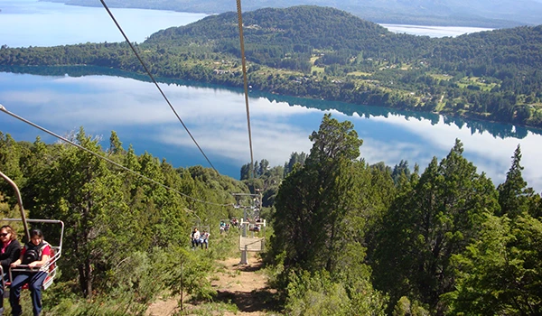 Paisagem natural de um lago rodeado por morros e vegetação verde, com céu claro ao fundo, capturada de um ponto elevado.