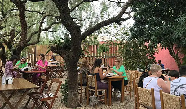Restaurante com ambiente aconchegante, contendo mesas ocupadas por pessoas. O local é bem iluminado e rodeado de vegetação. Ideal para refeições em grupo.