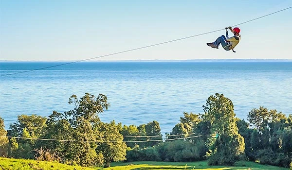 Pessoa se divertindo em tiroleza em um parque ao ar livre com vista para o mar e árvores verdes ao fundo, perfeito para lazer e aventura.