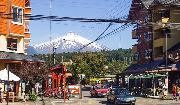 Vista de um vulcão ao fundo de uma rua movimentada em uma cidade com construções, carros e pessoas caminhando, mostrando o cenário urbano ao lado do símbolo da natureza na Travessia dos Lagos Andinos.