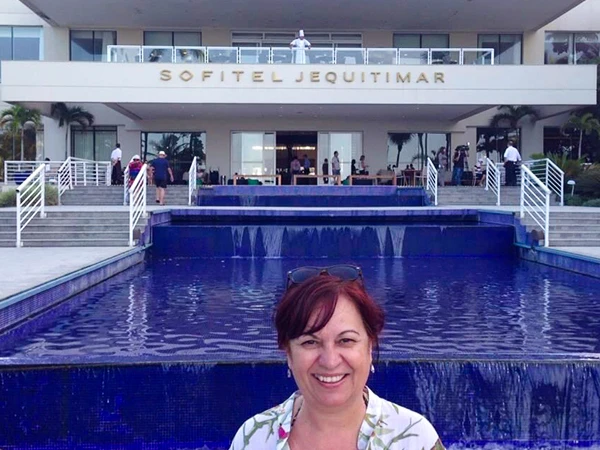 Deusa Rodrigues em frente ao hotel Sofitel Jequitimar, com fonte azul ao fundo. Ambiente acolhedor e elegante, ideal para relaxar.