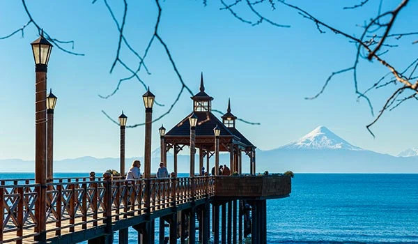Vista do píer de madeira com várias pessoas caminhando, iluminação de postes antigos, ao fundo o mar com vista para vulcão. Travessia dos Lagos Andinos.