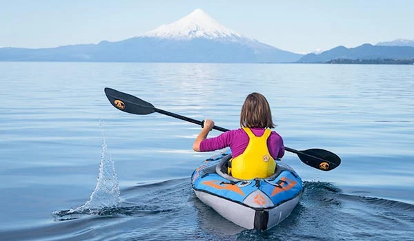 Pessoa jovem navegando de caiaque em um lago com vista para uma montanha coberta de neve ao fundo, no dia ensolarado. Travessia dos Lagos Andinos.