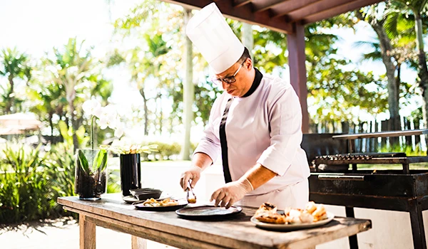 Chef preparando pratos em ambiente ao ar livre, utilizando ingredientes frescos e técnicas culinárias aprimoradas, destacando a gastronomia de qualidade.