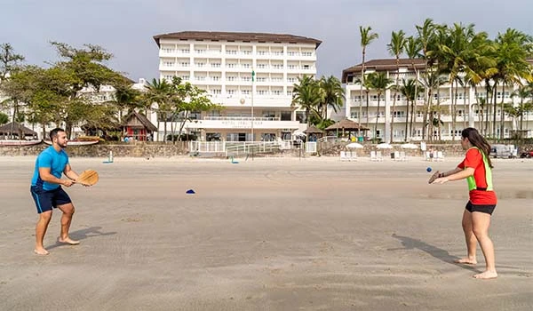Casal jogando tênis de praia na areia, com um hotel ao fundo e palmeiras nas laterais, proporcionando um dia ensolarado e agradável.