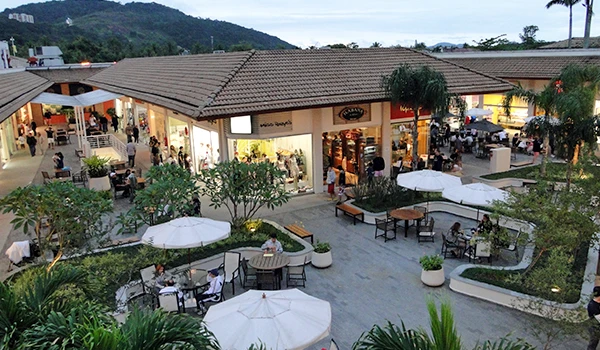 Vista aérea de um shopping ao ar livre com lojas, mesas e cadeiras, rodeado por áreas verdes, perfeito para compras e lazer.