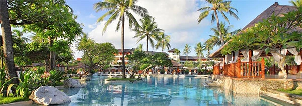 Imagem de uma piscina resort tropical cercada por palmeiras e cabanas de madeira, com céu azul claro e ambientes de lazer ao ar livre, ideal para férias e relaxamento.
