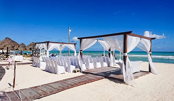 Estrutura de cerimônia na praia, com cadeiras brancas e gazebos de madeira com cortinas brancas, em dia ensolarado na orla. Casamento e renovação de votos no exterior.
