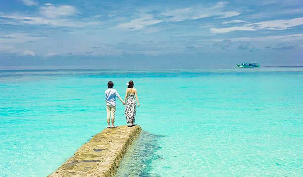 Casal caminhando de mãos dadas em um píer de pedra sobre águas cristalinas e azuis do mar em uma tarde ensolarada na praia tropical.