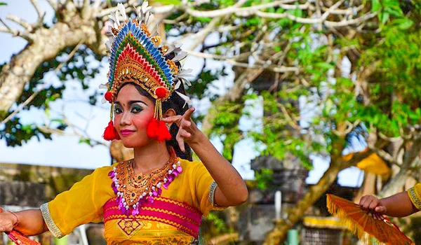 Mulher com traje tradicional colorido, com destaque para sua cabeça ornamentada, participando de uma celebração cultural ao ar livre em uma área com árvores e céu azul na viagem pela Ásia.