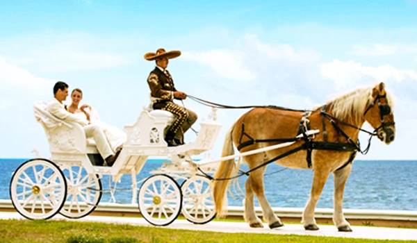 Cena de um carruagem puxada por um cavalo com uma pessoa vestida de mexicano tradicionalmente, ao lado de um casal sentado na carruagem, na orla marítima. Casamento e renovação de votos no exterior.