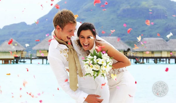 Casal celebrando casamento na praia, com celebração ao ar livre, confetes coloridos e cenário de lago e montanhas ao fundo.