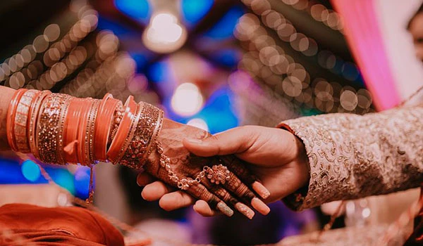Casamento tradicional com troca de alianças na cerimônia, destaque para mãos entrelaçadas e decoração festiva ao fundo.