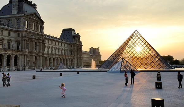 Vista do Museu do Louvre com a pirâmide de vidro ao entardecer em Paris, França, mostrando turistas caminhando e apreciando a arquitetura impressionante.