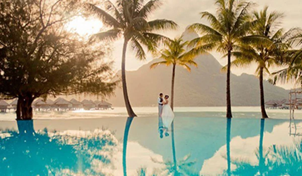 Casal de noivos dançando na beira de uma piscina, com cenário de pôr do sol, palmeiras e montanhas ao fundo, criando uma atmosfera romântica e tropical.