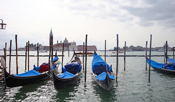 Gôndolas tradicionais de Veneza ancoradas na água, com o cenário de Veneza ao fundo e céu nublado. Uma cena típica da cidade italiana, famosa por seus canais e transporte de gôndolas. 