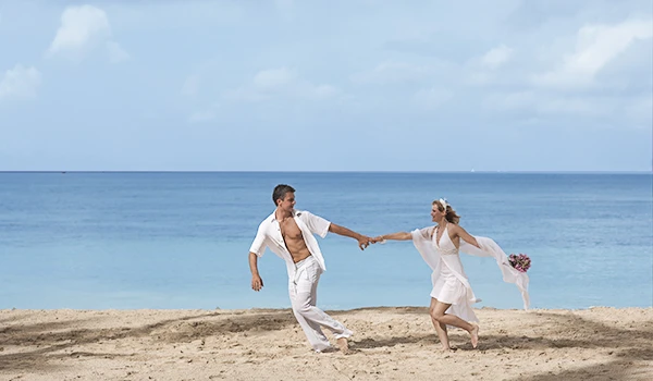 Casal de noivos brincando na praia, com céu azul e mar ao fundo, vestido de noiva e terno claro, demonstrando felicidade e amor em uma cerimônia ao ar livre na areia. Casamento e renovação de votos no exterior.