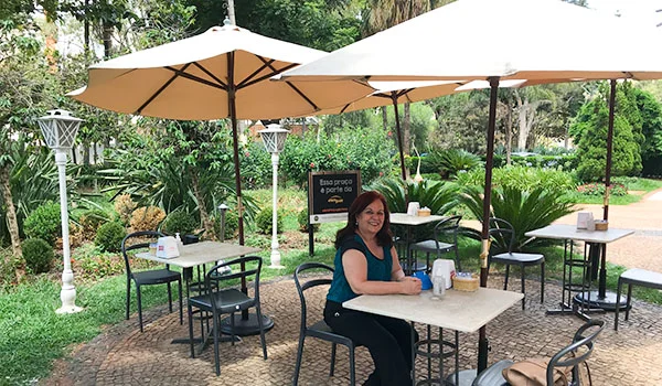 Foto de Deusa Rodrigues sentada em uma das mesas com guarda-sol ao ar livre em meio a jardins.