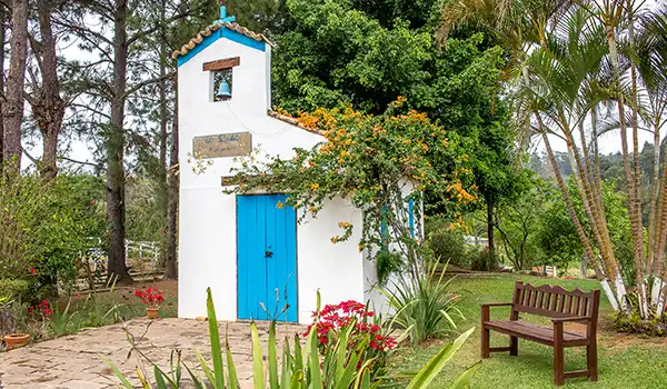 Uma linda capela branca com detalhes em azul, cercada por jardins floridos e árvores no campo, ideal para fotos e passeios ao ar livre.