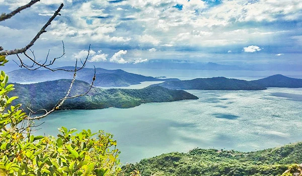 Vista panorâmica de um grande lago cercado por montanhas verdes sob céu parcialmente nublado, ideal para quem busca natureza e tranquilidade.