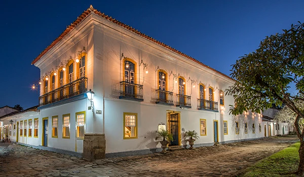 Hotel com fachada colonial de noite com janelas iluminadas e árvores ao redor, localizada em uma rua tranquila.
