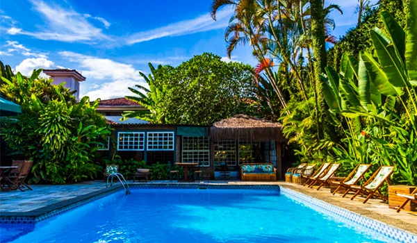 Imagem de uma piscina externa cercada por árvores em uma pousada, com céu azul e nuvens brancas ao fundo, ideal para relaxar e aproveitar o verão.