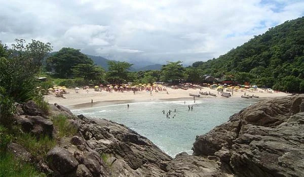 Praia com águas transparentes, cercada por árvores e montanhas ao fundo, com pessoas aproveitando o ambiente em um dia ensolarado na natureza.