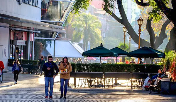 Pessoas caminham por uma rua arborizada, com mesas, cadeiras e guarda-sóis em uma área externa de cafés, em um dia ensolarado no bairro da Recoleta, em Buenos Aires, Argentina.