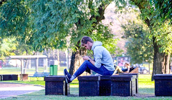 Homem sentado em um banco de praça, usando um celular, em meio a árvores frondosas e áreas verdes em um parque do bairro da Recoleta, em Buenos Aires, Argentina.