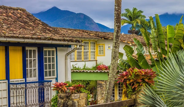 Casa colonial brasileira com varanda colorida, jardim exuberante com várias plantas tropicais, em cenário com montanhas ao fundo