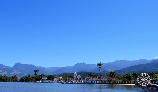 Praia de águas tranquilas com árvores, prédios e montanhas ao fundo, sob céu azul claro, cenário perfeito para relaxar e apreciar a natureza.