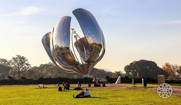 Escultura metálica gigante em formato de flor, instalada em um parque com gramado verde, árvores ao fundo e pessoas sentadas ao ar livre, sob a luz do pôr do sol.