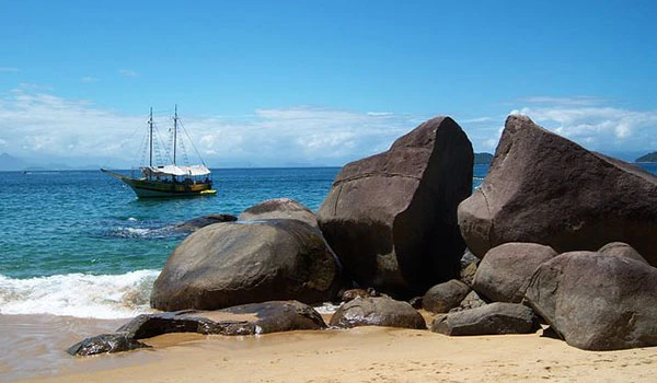 Cenário de praia com rochas grandes e um navio ao fundo no mar, sob céu ensolarado, perfeito para turismo e atrações marítimas. Lua de Mel em Paraty.