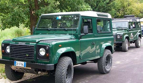 Imagem de vários veículos Land Rover Defender estacionados ao lado de uma área verde com árvores, destacando o modelo clássico e robusto, popular para aventuras fora de estrada.