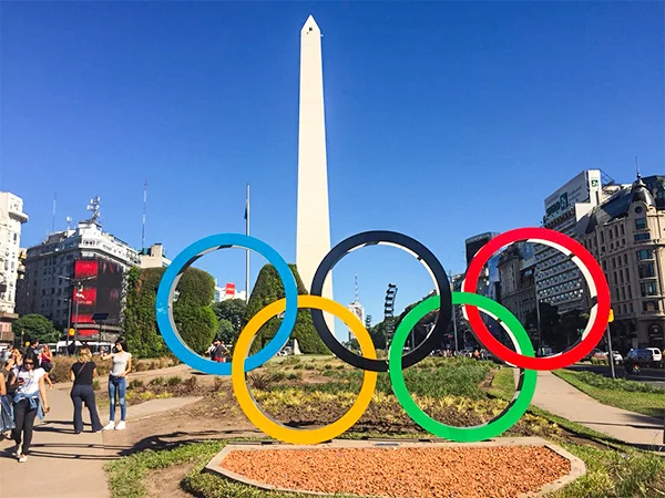 Monumento do Obelisco de Buenos Aires ao fundo e os cinco anéis coloridos do símbolo olímpico em primeiro plano, em uma praça ajardinada, sob um céu azul e cercados por edifícios e pedestres.