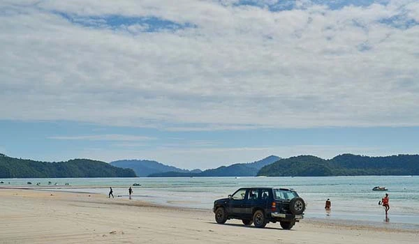 Imagem de uma praia tranquila com um carro na areia, pessoas caminhando na beira do mar e uma paisagem de montanhas ao fundo, sob céu com nuvens.