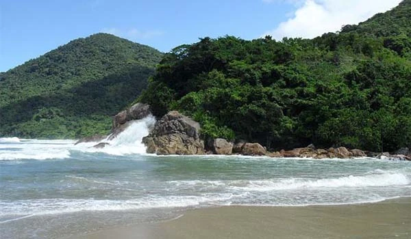 Praia deserta com ondas suaves, cercada por uma vegetação exuberante e uma serra ao fundo, em um dia claro e ensolarado.