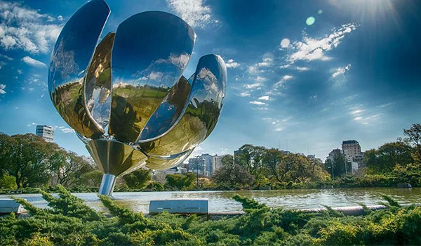 Escultura metálica em formato de flor, localizada em um parque às margens de um lago, sob um céu azul com nuvens brancas e sol intenso.