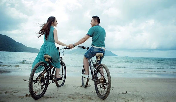 Casal de jovens apaixonados pedalando de bicicleta na praia sob céu nublado, apreciando o momento romântico na beira-mar, na Lua de Mel em Paraty.