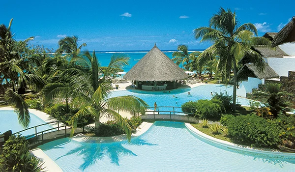 Foto do Belle Mare Plage Hotel com área de piscina tendo ao centro um bar molhado coberto de palha e cercado de coqueiros e mar. 