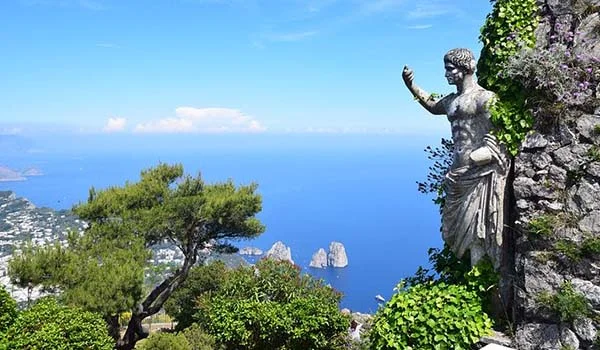 Vista panorâmica da ilha de Capri, na Itália, com uma estátua feminina junto a uma parede de pedra, vegetação ao redor e o mar azul ao fundo.
